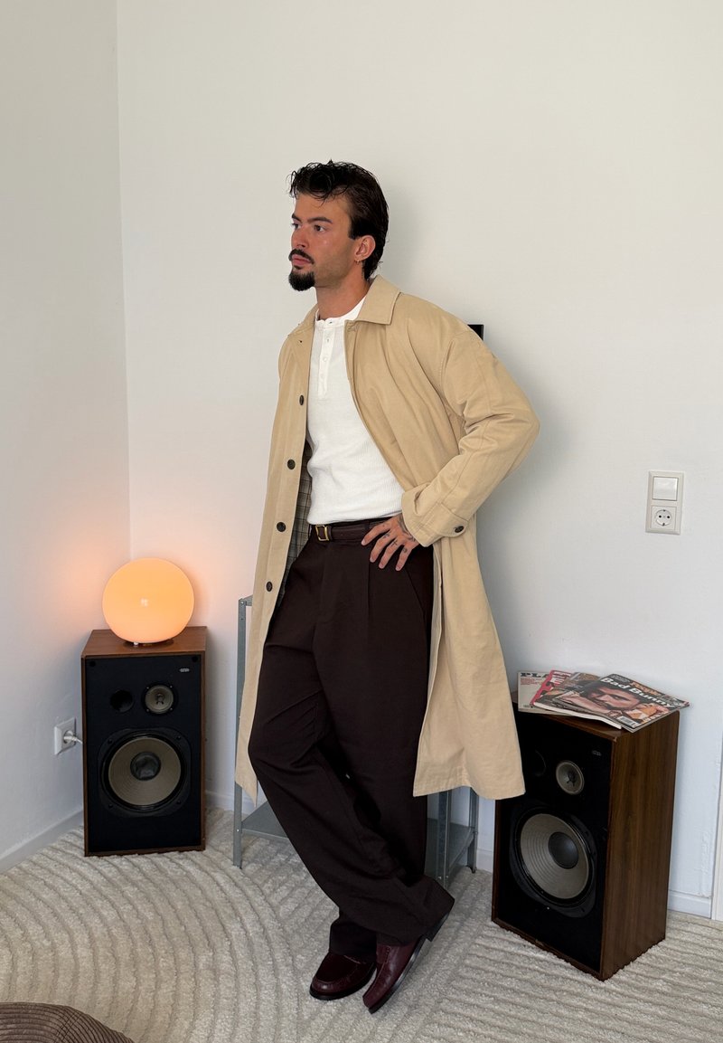 Homme barbu portant un manteau beige, une chemise blanche et un pantalon sombre, appuyé contre un mur entre deux enceintes dans une pièce minimaliste.