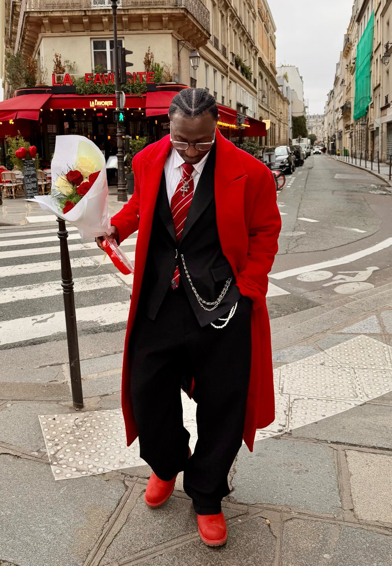 Homme en manteau rouge et chaussures tenant un bouquet de roses rouges et blanches dans une rue de Paris près d'un café aux auvents rouges.