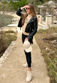 Černá kožená bunda, béžová mikina s grafikou, černé legíny, béžová kšiltovka a světlé tenisky s pastelovými akcenty na cestě u vody a zeleně.