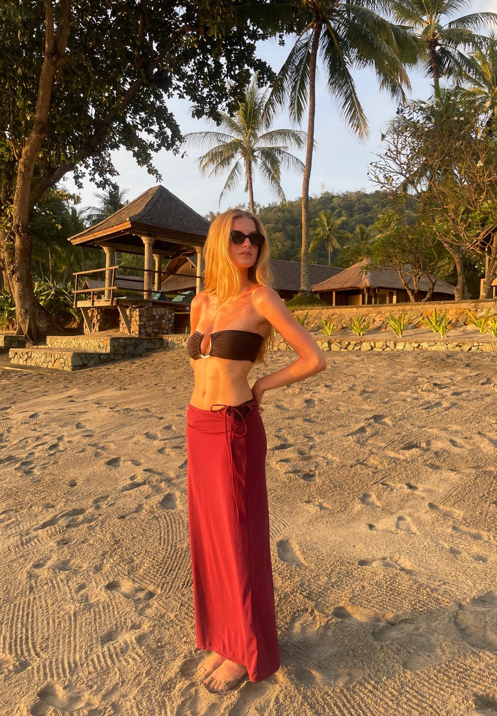 Černý bikiny top s klíčovou dírou a dlouhou, vínovou zavinovací sukni uvázanou v pase. Písečná pláž s palmami v pozadí.