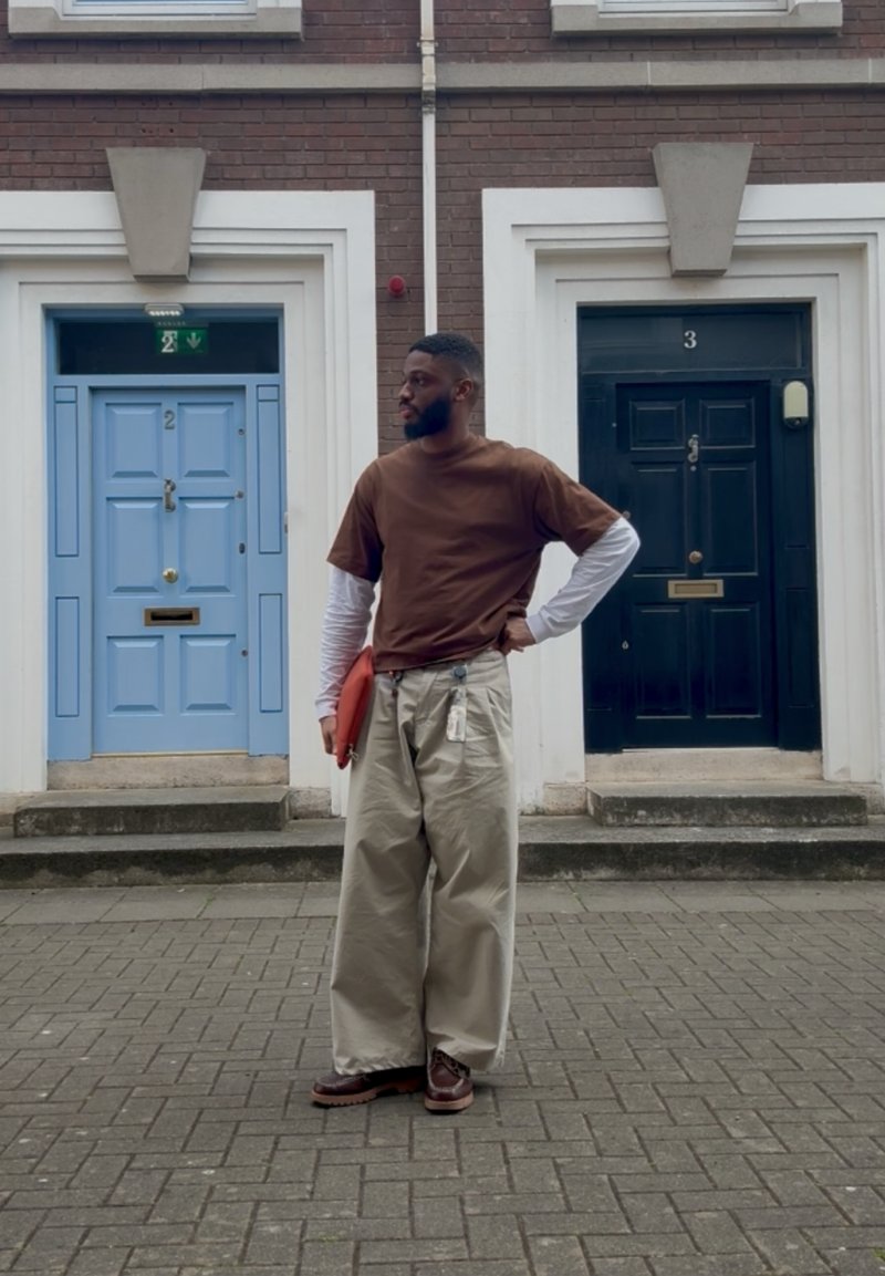 Homme en chemise marron et pantalon beige ample debout dans une rue pavée entre deux portes numérotées 2 et 3, tenant un dossier orange.