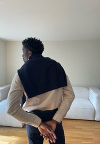 Bărbat cu păr creț și închis la culoare stă înăuntru cu mâinile încrucișate pe la spate, purtând o cămașă bej, pantaloni închiși la culoare și un pulover bleumarin așezat pe umeri.