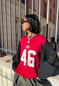 Rød NFL-drakt med hvitt nummer 46. Båret over en svart oversized jakke. Sølv kjede og solbriller. Bakgrunnen viser et gjerde.