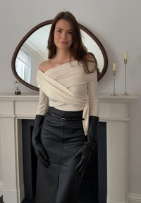 Γυναίκα με μακριά καστανά μαλλιά, που φοράει εκρού τοπ με ακάλυπτους τους ώμους, μαύρη δερμάτινη φούστα και μαύρα δερμάτινα γάντια, στέκεται δίπλα σε ένα λευκό τζάκι με κεριά.