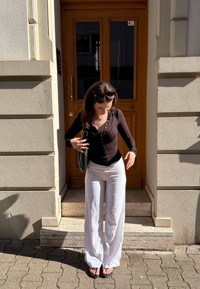 Mujer joven ajustando un top negro, lleva pantalones anchos blancos y chanclas, de pie al sol sobre escalones de piedra frente a una puerta de madera.