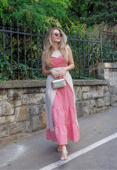 Mujer con vestido vichy rojo con sobreposición beige y gafas de sol, sosteniendo un pequeño bolso plateado, caminando por una acera cerca de un muro de piedra y vegetación.