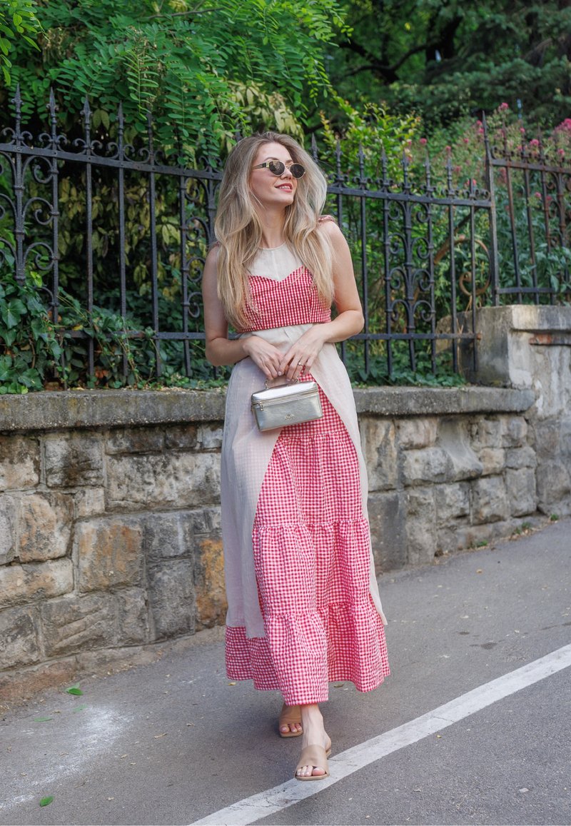 Femme portant une robe vichy rouge avec un dessus beige et des lunettes de soleil, tenant un petit sac argenté, marchant sur un trottoir près d'un mur en pierre et de la verdure.
