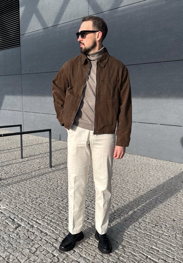 Brun fløjljakke over en beige turtle neck, parret med lyse bukser og sorte sko, sat mod en grå væg med mønstre.