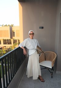 Lys grå oversized sweater, cremet maxiskirt og sorte flip-flops. Mønstret bælte, håndtaske i guldtonet og mørke solbriller. Rattan stol i nærheden.