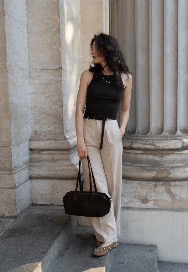 Zwarte tanktop, beige high-waisted broek en een bruine handtas met twee handvaten. Platte schoenen met decoratieve studs. Steenkolommen op de achtergrond.