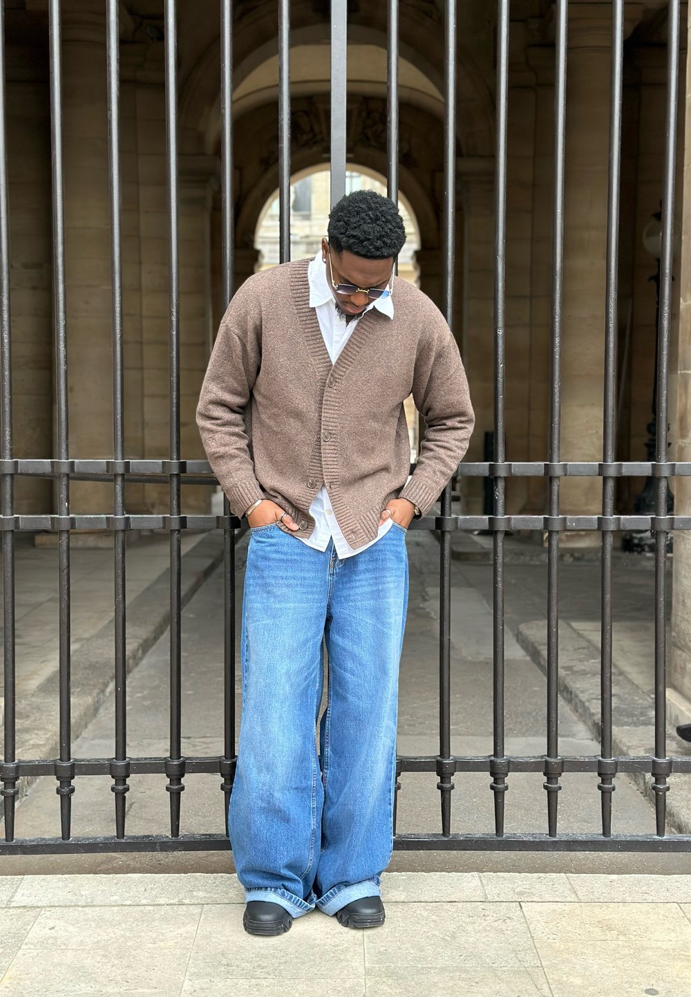 Brun cardigan, bílá košile a široké modré džíny. Stojí před černou kovovou bránou s vertikálními pruty. Neformální obuv.