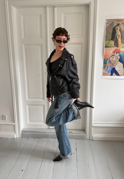 Persona con chaqueta de cuero negra, gafas de sol y vaqueros levanta una pierna mostrando botas negras puntiagudas, de pie frente a puertas blancas y arte en la pared.