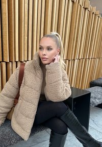 Beige faux fur zip-up jacka med hög krage, tillsammans med svarta leggings och knähöga blanka svarta stövlar, mot en bakgrund av träpanel.