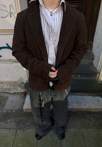 Blazer de camurça castanho com costuras em contraste, camisa listrada de botão, jeans cinzentos com correntes e sapatos pretos brilhantes. Acessorizado com anéis e um colar.