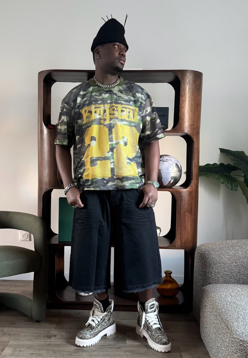 Homme debout à l'intérieur portant un t-shirt en mesh camouflage avec le numéro jaune "22", un short large noir, des bottes à motifs, un chapeau noir avec des pin's et un collier en chaîne argentée.