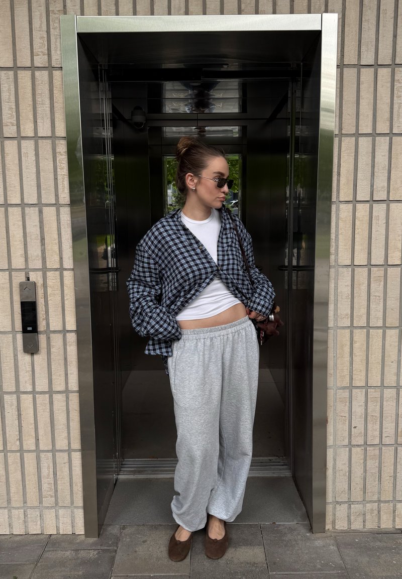 Jeune femme portant des lunettes de soleil, un pantalon de survêtement gris et une chemise à carreaux, debout à l'entrée de l'ascenseur dans un bâtiment aux murs carrelés.