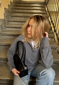 Grå bomullströja med knapppynt, denimjeans och en texturerad svart handväska, sittande på stensteg med ett räcke i bakgrunden.