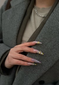 Lange, puntige nagels in transparant wit en paars met gouden en zilveren accenten. Hand rust op een grijze wollen jas, waarbij de textuur zichtbaar is.