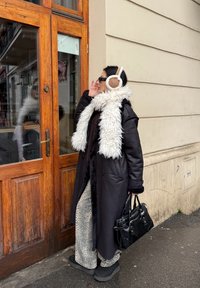 Lang sort frakke med faux fur kant, mønstrede bukser og sorte ankelstøvler. Hvide ørevarmere og en sort håndtaske fuldender looket.