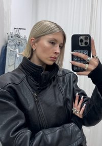 Jeune personne aux cheveux blonds courts vêtue d'une veste en cuir noire prenant un selfie dans un miroir en tenant un smartphone, avec des jeans accrochés en arrière-plan.