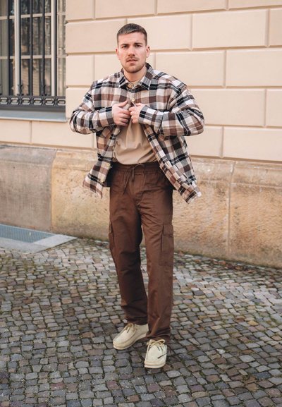 Camisa de cuadros en beige, marrón y negro sobre una camiseta color crema; pantalones cargo marrones con bolsillos; zapatos beige. De pie sobre una superficie de adoquines.