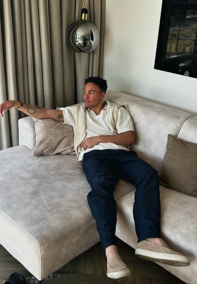 Sofá seccional tapizado en beige claro con superficie texturizada, pantalones navy y zapatos tipo slip-on beige. Se puede ver un cojín decorativo y una lámpara de pared.