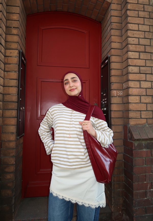 Vrouw met een wijnrode hijab en gestreept shirt staat zelfverzekerd en houdt een grote wijnrode tas vast voor een rode deur omgeven door bakstenen muren.