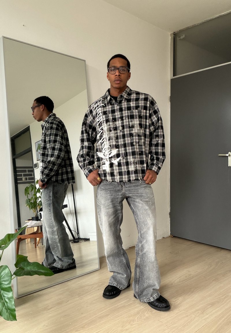 Mann med briller, svart- og hvit rutete skjorte med hvit tekst, grå slitne jeans og svarte sko står foran et helfigur-speil.