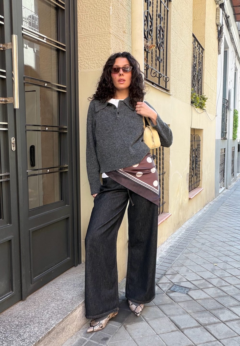 Femme aux cheveux bouclés portant des lunettes de soleil, un pull gris, un pantalon large noir, une ceinture écharpe à motifs, des chaussures en peau de serpent et un sac à main jaune dans une rue de la ville.