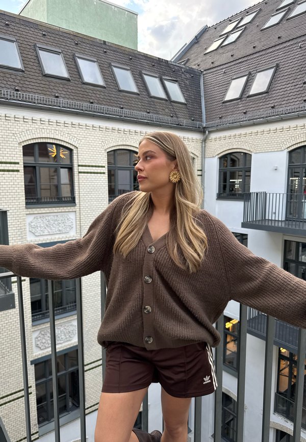 Bruine gebreide cardigan met grote knopen, gedragen over donkere bruine shorts met witte strepen, voor een patroon van een gebouw.