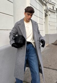 Casaco cinzento com padrão espinha de peixe e lapelas largas, luvas pretas, camisa branca e jeans azuis. Segura uma pequena bolsa preta. Ambiente urbano casual.