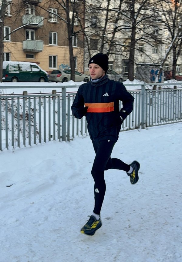 Uomo che corre su un sentiero innevato indossando abbigliamento sportivo nero, berretto nero e scarpe da corsa gialle vicino a una recinzione metallica in un contesto urbano invernale.