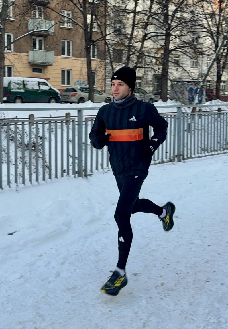 Homme courant sur un chemin enneigé portant des vêtements de sport noirs, un bonnet noir et des chaussures de course jaunes près d'une clôture métallique dans un cadre urbain hivernal.