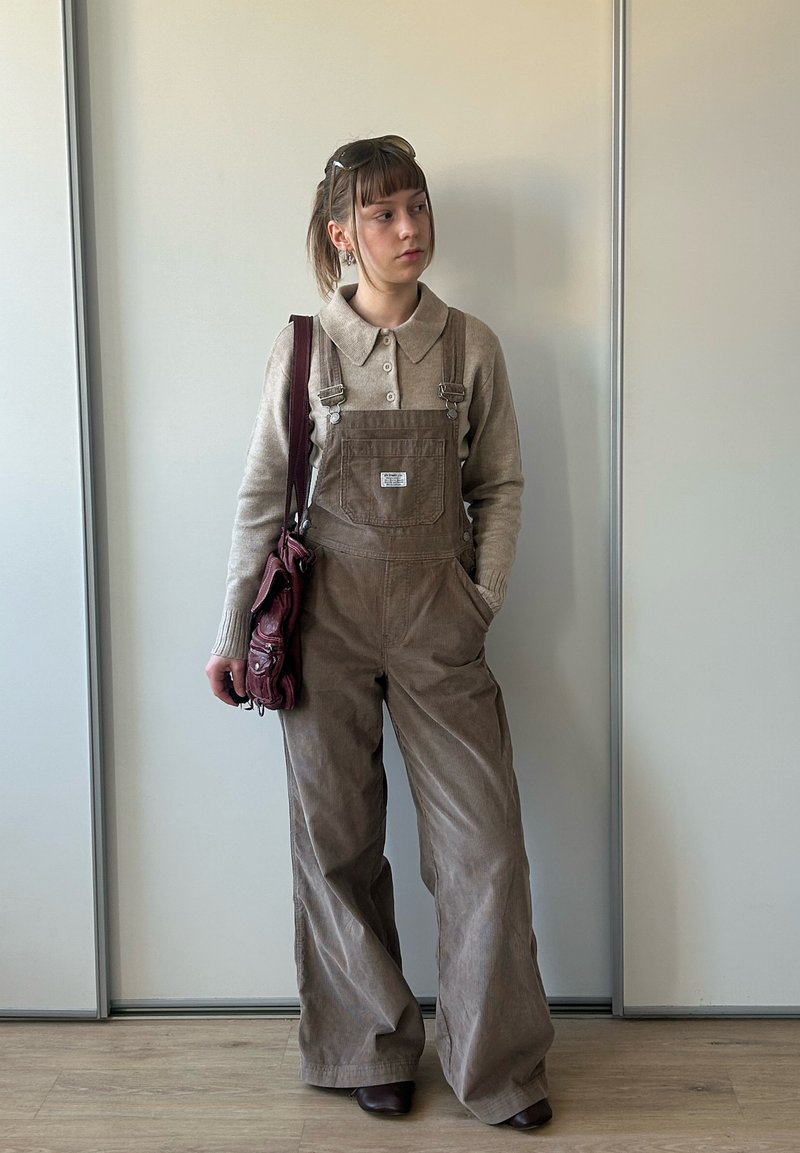 Jeune femme portant une salopette en velours côtelé marron et un pull beige, debout avec une main dans la poche et portant un sac à bandoulière bordeaux.
