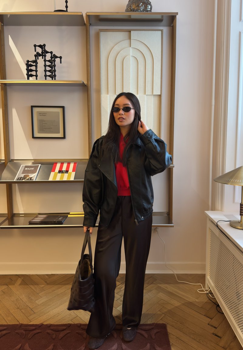 Woman wearing black leather jacket, red top, dark wide pants, sunglasses, holding black handbag indoors by shelves and window.