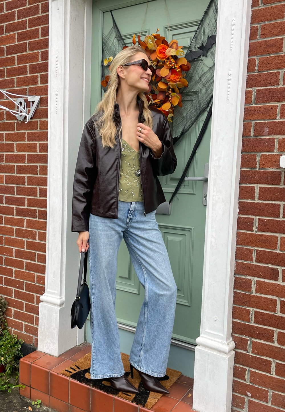 Brown leather jacket over a green top with beaded detailing, paired with wide-leg light blue jeans and black heeled boots, standing at a door.