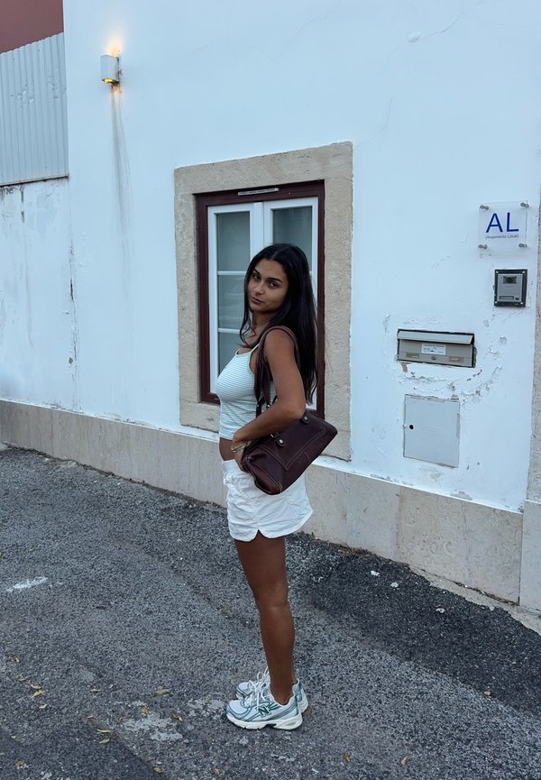 Mujer joven con el pelo largo y oscuro, lleva una camiseta sin mangas blanca, pantalones cortos blancos y zapatillas, y está de pie en la acera junto a un edificio blanco con una ventana.