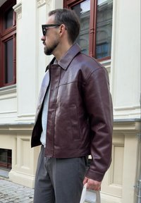 Bărbat îmbrăcat cu o jachetă de piele maro închis, pantaloni gri și ochelari de soare, ținând o geantă albă de tip tote, stând lângă o clădire de culoare crem cu feronerie.