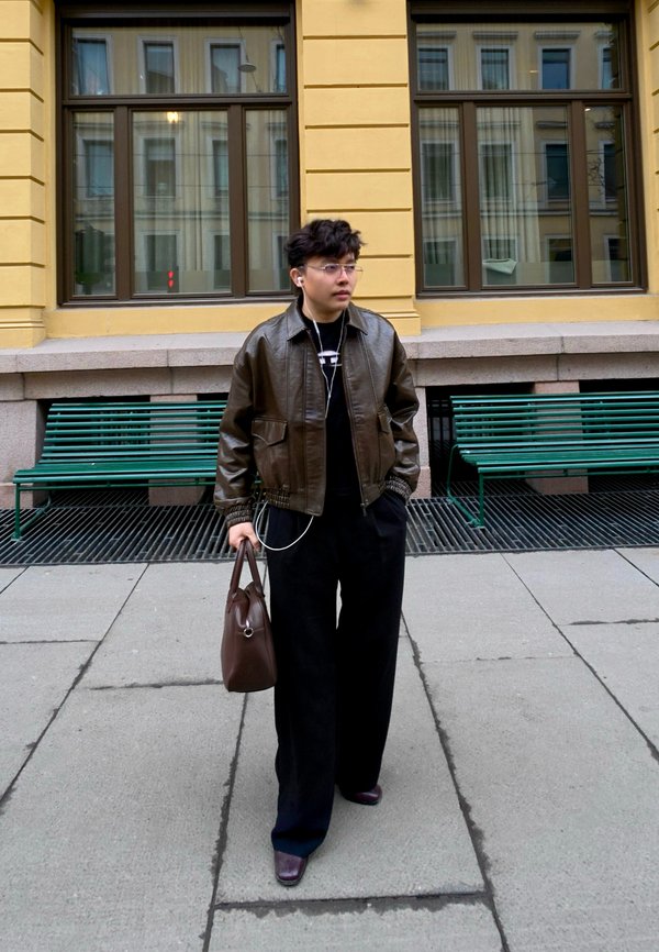 Junge Person mit kurzen Haaren trägt eine braune Lederjacke, schwarze Hose, trägt eine braune Tasche und läuft auf dem Bürgersteig der Stadt in der Nähe grüner Bänke.