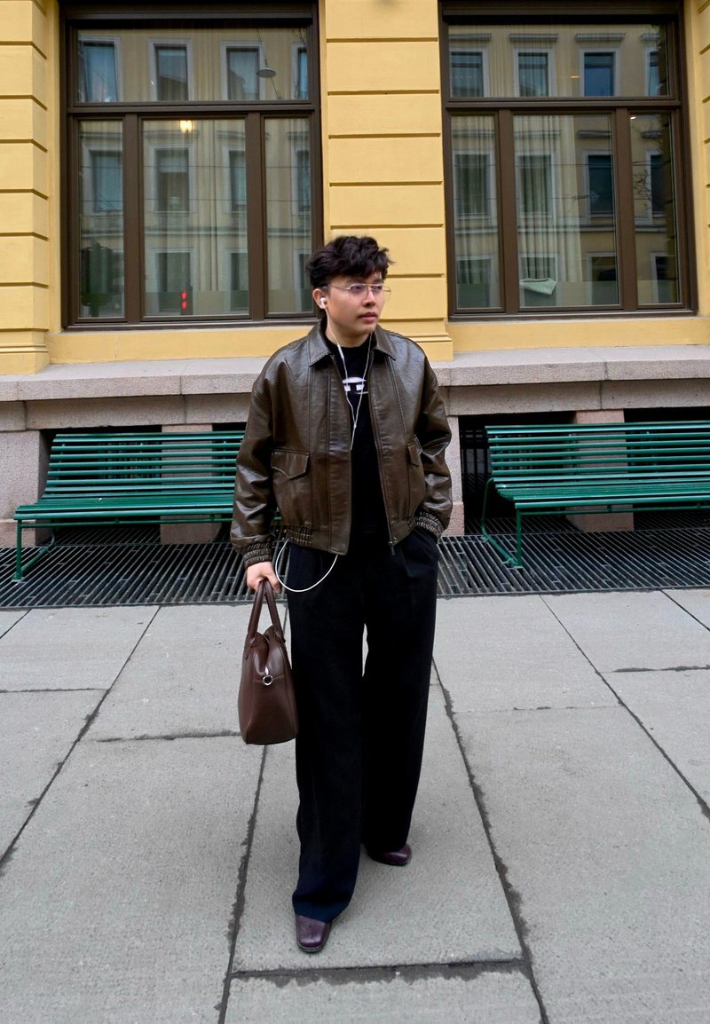 Jeune personne aux cheveux courts porte une veste en cuir marron, un pantalon noir, porte un sac marron, marche sur le trottoir de la ville près de bancs verts.