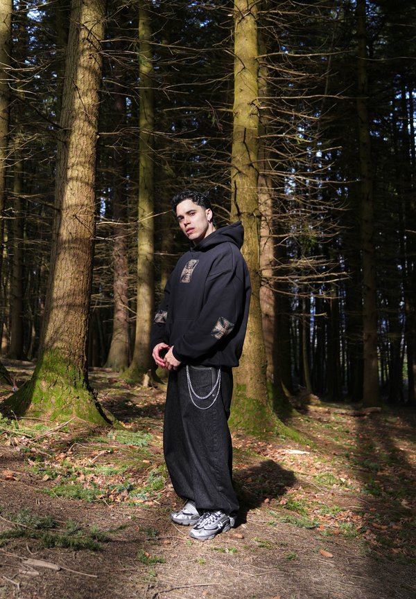 Ung mann i svart hettegenser og baggy jeans med kjede står i en solfylt skog med høye trær og spredte blader.