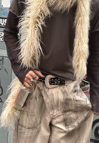 Loose beige trousers with visible stitching, a black belt adorned with rhinestones, and a textured white faux fur scarf. A hand holding a drink.