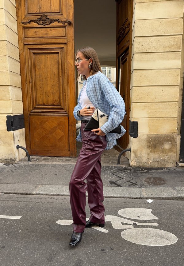 Une femme en chemise à carreaux bleus et pantalon bordeaux sort par une porte ornée en tenant une pochette noire et blanche.