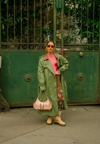Femme portant un trench-coat vert, un pull rose, des collants à losanges, des bottes beige, tenant un sac à main rose, debout devant une porte en métal verte.