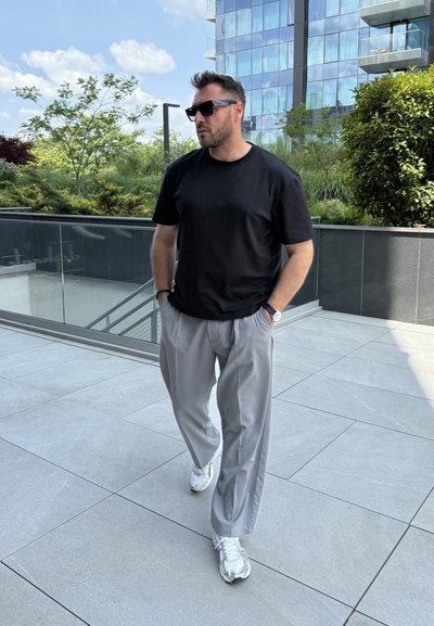 Una camiseta negra de manga corta, pantalones grises claros de corte holgado y zapatillas deportivas plateadas. El fondo presenta vegetación y arquitectura moderna.