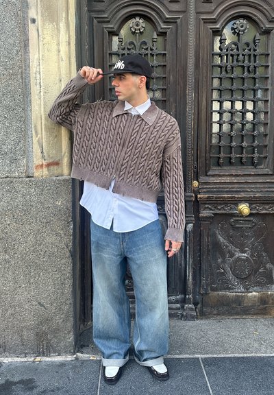 Suéter corto marrón tejido a crochet, camisa blanca con cuello, jeans anchos azul claro, gorra negra y zapatos negros con detalles en blanco.