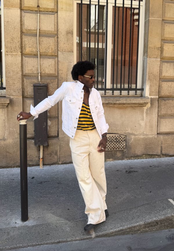 Personne portant une veste blanche et un pantalon crème s'appuie sur un poteau noir sur un trottoir de la ville avec un bâtiment en pierre beige et une fenêtre grillagée en arrière-plan.