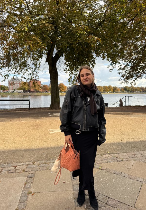Sort læder cropped bomberjakke, sort lang kjole, mønstrede sorte ankelstøvler, vævet orange håndtaske, grå strikscarf, naturbaggrund.