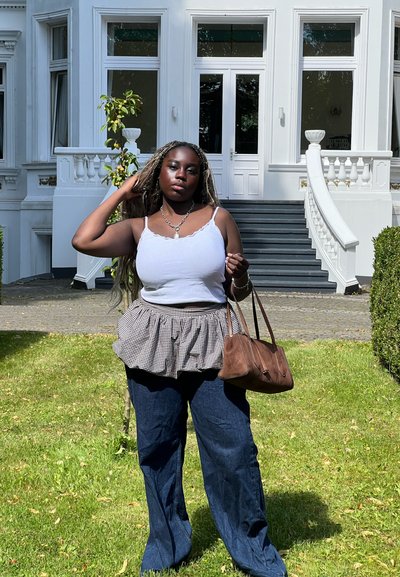 Top blanco sin mangas, falda peplum a cuadros en blanco y negro, jeans de campana azul oscuro, llevando un bolso marrón. Fondo de césped verde y edificio blanco.