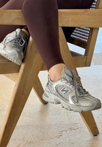 Grey athletic shoes made of mesh and synthetic materials, complete with round laces and a textured sole, placed against the leg of a wooden chair.